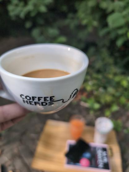 White coffee cup with 'coffee nerd' written on it. Vaguely in the background: a tray with some other breakfast items, a book and a phone, ajd some green bushes