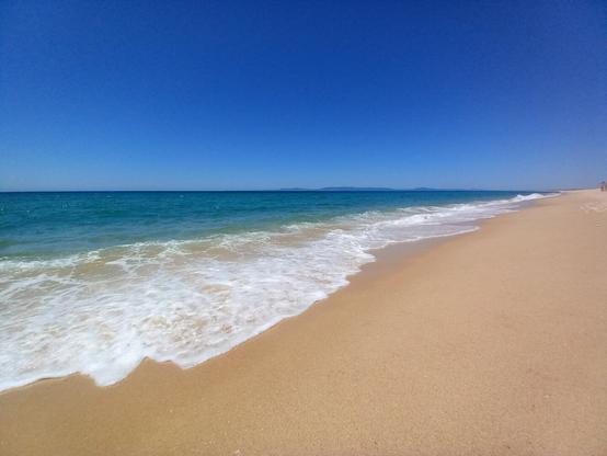 A pristine sandy beach with gentle waves lapping the shore under a clear blue sky; the sea transitions from turquoise near the shore to deep blue towards the horizon, with distant hills faintly visible.
