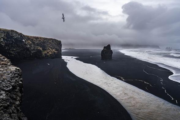 View of Reynisfjara Beach in Iceland, featuring the sea stack Arnardrangur in the foreground and the Reynisdrangar basalt formations in the distance under a moody, overcast sky. The dark volcanic sand and dramatic atmosphere reflect the rugged Icelandic coastline.