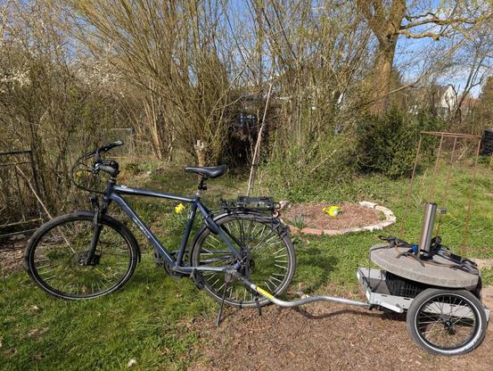 Fahrrad mit Anhänger auf dem sich ein schwerer Sonnenschirmständer befindet. Das Rad steht in einem Garten