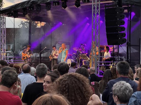 Eine Live-Musikaufführung von Dota und ihrer Band, aufgenommen bei einer Veranstaltung im Freien. Auf der Bühne gibt es eine Band mit mehreren Musikern, darunter eine Sängerin und Gitarristen, die von lila Bühnenlichtern beleuchtet werden. Eine Menge Zuschauer versammelt sich vor der Bühne und genießt die Aufführung.
