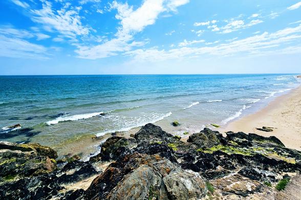 A wide shot of a sunny beach scene with a clear blue sky and fluffy white clouds. In the foreground, dark, rugged rocks covered with some green moss extend into the gentle waves of the turquoise sea. The middle ground features the calm ocean with small white-capped waves rolling towards a wide, golden sandy beach that stretches into the distance on the right. The overall mood is peaceful and inviting.