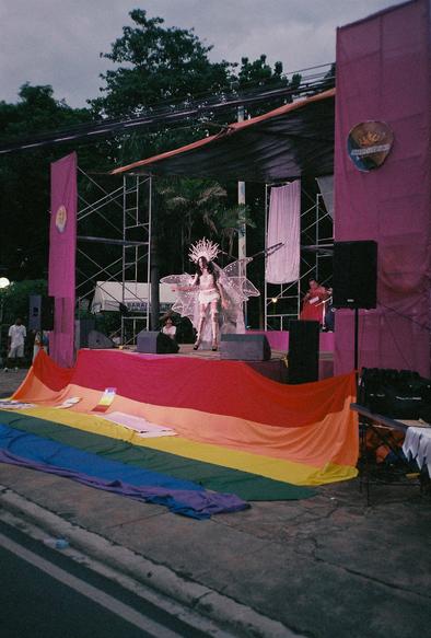 A stage with a drag performer in a white winged costume and fan-like headdress. There is a giant rainbow flag tied to the stage and lying on the ground.