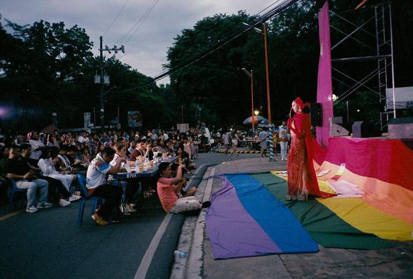 A photo from a Pride pageant: left side of the photo captures the audience and the judges, on the right side is a drag performer in an elegant red gown posing in front of a giant rainbow flag.