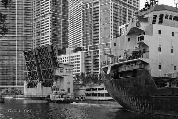 This is a black and white nautical / cityscape photo in landscape format. Between the right margin and the centre of the frame is the stern of a cargo ship. To the left of the ship is a tug attached to the ship with two stout lines. The ship is being towed backwards along a city river from right to left. Our ship fills nearly the entire right side of the image. The very weathered and battered hull is black while the superstructure over the stern is white. The tug is approaching a raised bascule bridge. Surrounding the bridge are a number of very high-rise modern apartment buildings whose top floors are out of view. 
The Location: The Miami River In downtown Miami. The Raised Bridge is the Brickell Ave Bridge (2012).