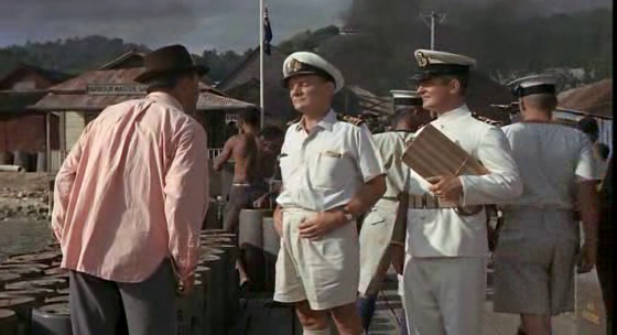 La imagen muestra una escena al aire libre en lo que parece ser un puerto o muelle en un clima tropical. Tres hombres son el punto focal de la imagen. Uno lleva un sombrero fedora y una camisa rosa, otro lleva un uniforme de capitán de la marina blanca, y el tercero lleva un uniforme similar pero con un portapapeles.  Al fondo se ven edificios de madera, humo, y varios hombres en ropa de trabajo o casual. El ambiente sugiere una escena de una película o un evento histórico.