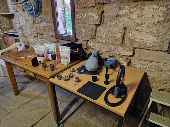 In einem alten Schuppen stehen zwei Tische. Darauf liegt diverses Kaffee-Equipment. Dazwischen sitzt ein kleiner Plüschhai.