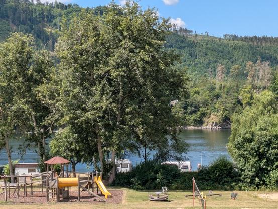 Im Vordergrund absteigend zu einer Wasserfläche ein Spielplatzgelände. Dahinter Hecken und Bäume vor dem Zugang zur Badestelle. Die Wasserfläche wird gegenüber von einer bewaldeten Berghöhe begrenzt.