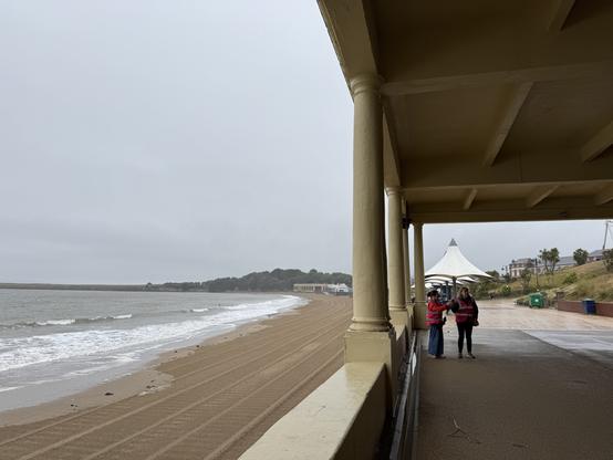 View from a partly covered seaside promenade. The beach to the left is empty, the sky and sea are both a dull grey and it’s raining.