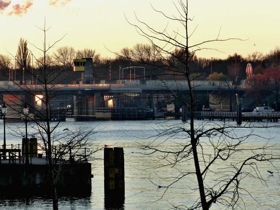 A calm river scene at sunset, featuring a bridge with a yellow control tower, bare trees in the foreground, and the reflective glow of the setting sun on the water and metal surfaces. A sculpture and a few birds add visual interest.