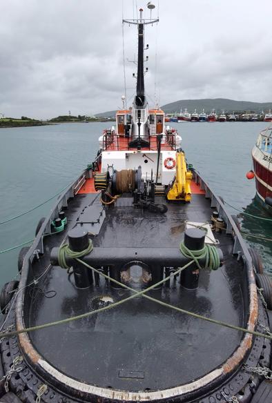 Stern view of the tug described in the text, seen from the pier it is moored to. The camera angle emphasizes the working deck's pronounced ellipsoidal shape. A very large towing bitt is visible and behind it the towing winches and a small folded hydraulic crane. In the distance, the deck house and mast.