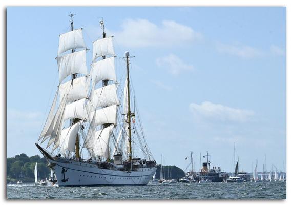 Das Foto zeigt das deutsche Segelschulschiff Gorch Fock während der Windjammerparade zur Kieler Woche 2025. Die Gorch Fock befindet sich im Vordergrund des Bildes und läuft mit gesetzten weißen Segeln an der Heikendorfer Bucht vorbei. Das stolze, dreimastige Segelschiff ist in voller Fahrt und dominiert das Bild mit seinen hoch aufragenden Masten und strahlend weißen Rahsegeln, die sich im Wind wölben.

Der Schiffsrumpf ist weiß mit einem schwarzen Ankerzeichen am Bug. An Deck sind zahlreiche Menschen zu erkennen – wahrscheinlich Kadetten, Besatzung und Gäste. Die Takelage ist komplex und deutlich sichtbar.

Im Hintergrund tummeln sich viele kleinere und mittlere Boote sowie einige weitere größere Schiffe, die ebenfalls an der Parade teilnehmen. Am Horizont ist die Küste mit grünen Bäumen zu sehen, der Himmel ist hellblau mit ein paar leichten, weißen Wolken. Die Wasseroberfläche ist leicht bewegt und reflektiert das Sonnenlicht.