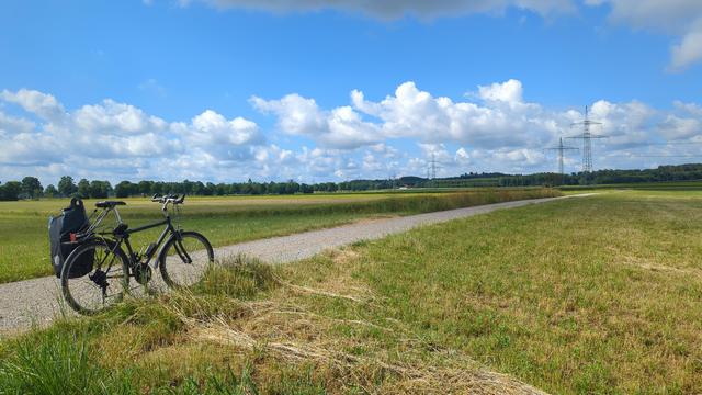 Bike with some carelessly mounted luggage on small road in between green fields and beneath blue sky with clouds.