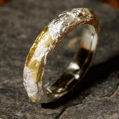 A close-up of a unique ring featuring a textured design with a mix of silver and gold finishes, resting on a dark stone surface.