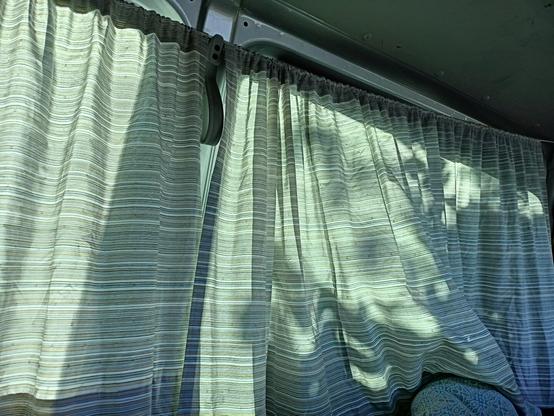 We see a curtain in a van where the shadowlines of the leaves on the other side of the curtain are outlined by the sunlight. The curtains are designed in white, grey and beige stripes.