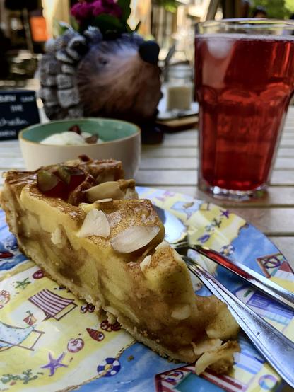 Auf einem kleinen Holztisch stehen im Vordergrund ein Teller mit Apfelkuchen, Vanilleeis sowie eine hausgemachte himbeer-Aronia-Limonade.