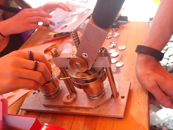Eine Buttonmaschine auf einem Tisch und Hände, die diese gerade benutzen.