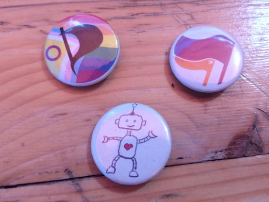 3 Button, einen mit Progressiv-Pride-Flagge und PiratenhP, eine mit lila und orangener Flagge und einer mit selbst gemalten Roboter.