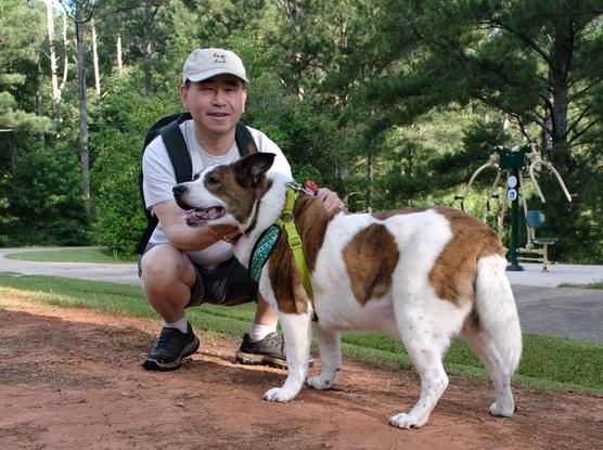 A man with a white cap squatting by a park trial holding his white and brown dog