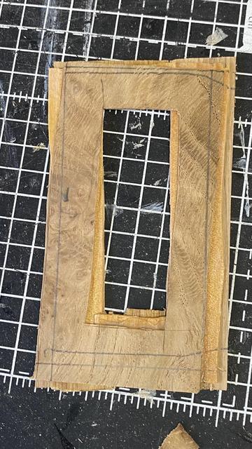 A sandwich of 3 wood veneers with a rough rectangular shape cut out, resting on a gridded cutting mat. The wood shows natural grain patterns and has pencil lines showing how it might be trimmed. It lies on a well-used black and white cutting mat.