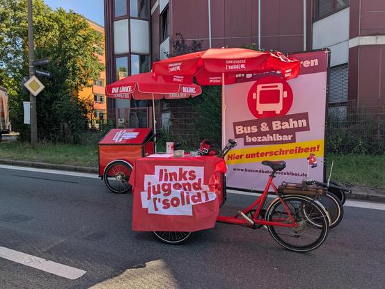 Zwei rote Lastenrader mit Sonnenschirmen von der Partei die Linke und deren Jugendorganisation Linksjugend ['solid]. Ein Lastenrad zieht einen Anhänger mit Plakat vom Bürgerbegehren Bus und Bahn bezahlbar. an dem anderen Lastenrad hängt eine Linksjugend ['solid] und eine kleine Kuba Fahne.