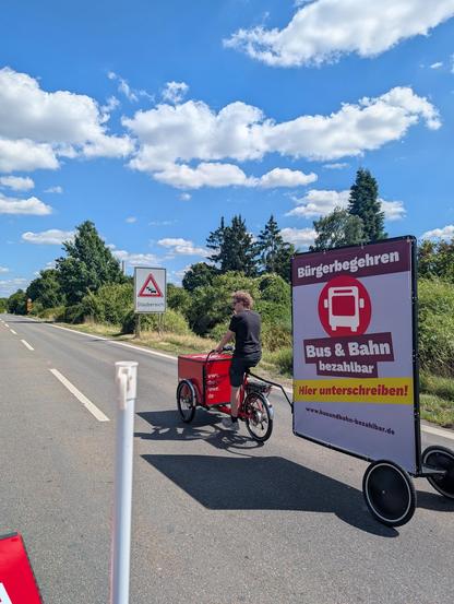 Ein rotes Lastenrad fährt auf dem Frankenschnellweg mit einem Plakatanhänger. Auf dem Plakat wird für das Bürgerbegehren Bus und Bahn bezahlbar geworben. Im Hintergrund ist ein großes Schild zu sehen, dass vor der Staugefahr dort warnt.