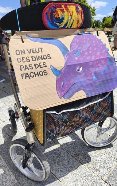 Sur un rollator décoré d'une broderie rainbow et d'un sac a rayures multicolores est fixée une pancarte en carton. Dessus un tricératops l'air fâché avec une corne cassée, il est aux couleurs du drapeau bi (bleu violet rose). A côté de lui est écrit en noir "On veut des dinos pas des fachos"