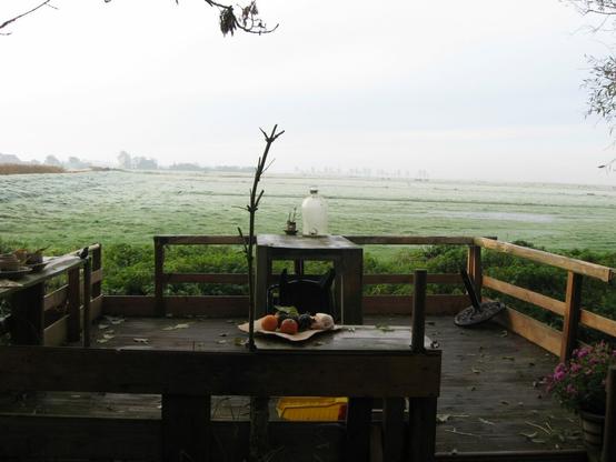 A misty rural landscape viewed from a rustic wooden terrace. On the table, a jug and some fruit add a touch of still life. The open field stretches into the distance under a pale, overcast sky.
