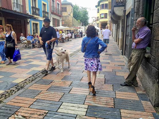 Un paisano fumando un pito pensativo, apoyado en la ventana de una taberna marinera, La Bodega de Rivero, en Avilés. Leva unos tirantes con la bandera tricolor de la segunda república. Abundante gente sentada en terrazas y caminando, en una bonita calle peatonal, con casas de no más de tres plantas con variedad de colores similares a los de las barcas.