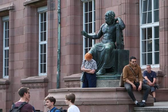 Große Bronzestatue einer antiken Persönlichkeit mit nach vorne gestrecktem Arm und Hand, der Unterkörper von einer Tunika bedeckt; unter der Hand der Statue eine Frau mit rotem Haar und gesenktem Kopf; rechts zwei sitzende Männer, auch zu Füßen der Statue; links drei junge Männer, von denen nur Kopf und Schultern zu sehen sind. / A large bronze statue of an ancient personage with one arm and hand stretched forward, the lower part of the body covered by a tunic; under the statue's hand, a woman with red hair and bowed head; to the right, two seated men, also at the statue's feet; to the left, three young men, of whom only the head and shoulders are visible.