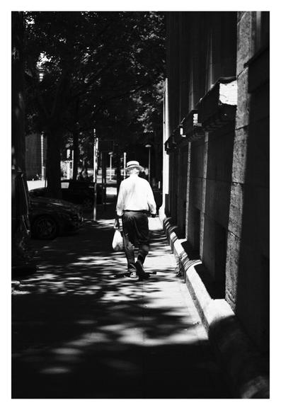 Schwarz-weiß Foto im Hochformat. Blick entlang eines Gehweges, rechts ist die Wand eines Gebäudes, links sind Bäume. Fast alles liegt im Schatten, nur mittig scheint die Sonne hin. Genau an dieser Stelle läuft eine Person von der Kamera weg. Dunkle Hose, weißes Hemd, weißer Sonnenhut. Die Person trägt in der linken Hand eine Tüte. Das gesamte Bild ist sehr kontrastreich.