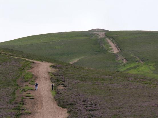 The broad path up 2,334ft Tinto. The image shows a view up the upper part of a bulbous mountain tp a wide cairn on its summit. A broad pink path leads up the mountain, over an intermediate rise. There are two people walking up the path, though one is each side of it. The light is subdued.