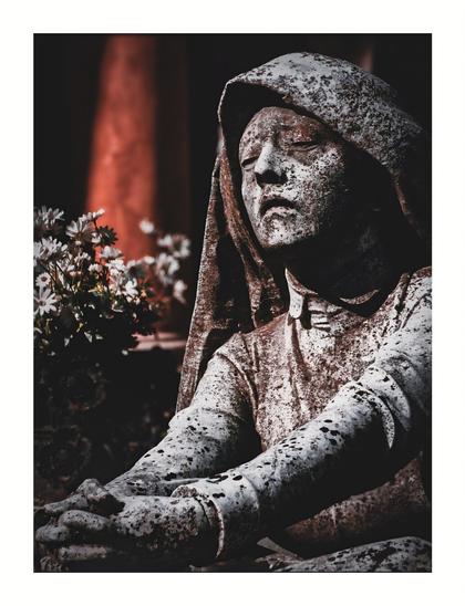 This photo is of a weathered stone statue of a hooded woman with her hands clasped together.  The statue is outdoors - with a blurry background and some white flowers visible in the foreground/midground. The colour palette is muted and dark. There's a reddish-brown tone mostly in the background and the statue itself shows signs of significant age and erosion.