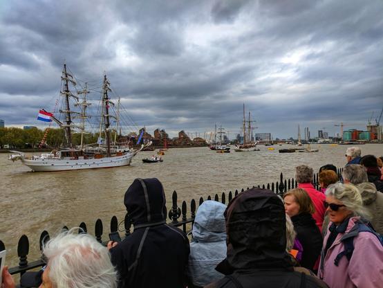 A large, three-masted tall ship, flying a Dutch flag from its stern, sails down a wide, muddy river under a heavily overcast sky. A small tugboat is beside it. In the background, other tall ships are visible further down the river, and the cityscape includes various buildings and industrial cranes. In the foreground, from a slightly elevated position, a crowd of people with their backs to the viewer are watching the ships. They are standing behind a black metal fence with decorative finials. Some people are wearing hooded jackets, and one person on the far right has light-coloured hair and is wearing a pink jacket.