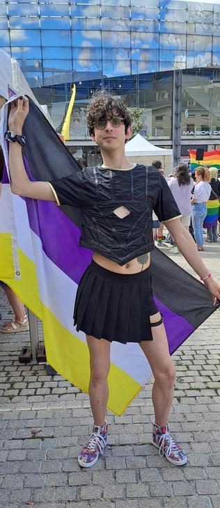 Photo de moi à la pride. Je tiens un drapeau non-binaire derrière moi, de travers. Je porte un jupe noire et un haut cousu sur mesure. C'est une sorte de crop top noir avec le bas qui est croppc en pointe. Au niveau de ma poitrine, il y a un trou en losange. Au cou et au manches, les bords sont dorés.