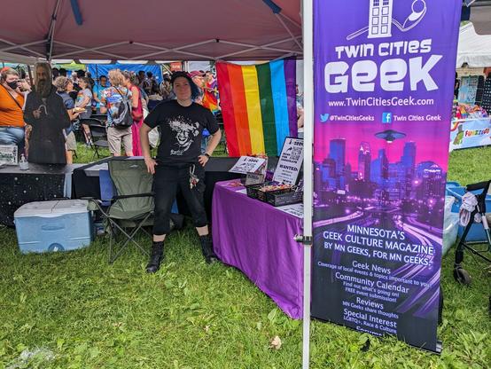 A woman with blue hair wearing a T-shirt with Marsha P. Johnson's face stands with her hands on her hips next to a Twin Cities Geek banner and table