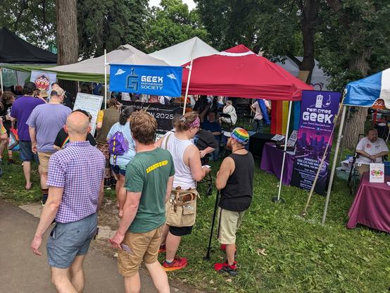 People pass by and stand near a red tent standing on grass with banners for Geek Partnership Society, Gaylaxicon, and Twin Cities Geek