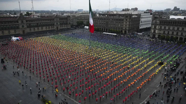 The image shows an impressive, coordinated demonstration in Mexico City’s Zócalo, the historic central square. Hundreds of people are standing in precisely organized rows, each holding a colored umbrella above their head to collectively form a giant rainbow flag. The colors span from left to right in the traditional rainbow order: red, orange, yellow, green, blue, and purple. In the center of the square stands a massive Mexican flag on a tall flagpole.
