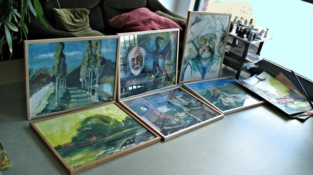 A collection of framed paintings is displayed on the floor near a sofa and window. The artworks include expressive portraits, landscapes, and abstract scenes. A nearby table holds small sculptures or chess pieces, adding to the creative, studio-like atmosphere.