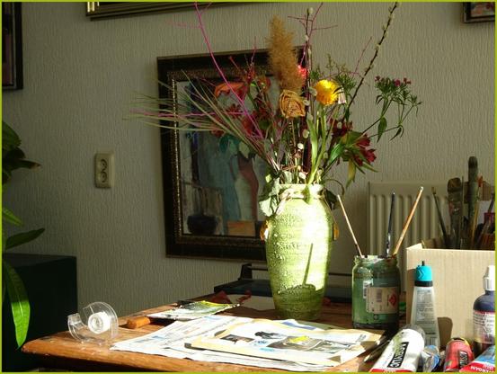 A sunlit artist’s workspace with a green vase holding a wild, colorful bouquet of dried and fresh flowers. The table is cluttered with painting tools—brushes, tubes of acrylic, tape, and newspapers. In the background, framed artwork hangs on a softly textured wall.