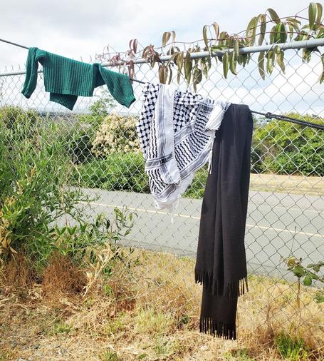 A medium shot captures a gray chain-link fence running horizontally across the frame. Over the fence, at different points, hang three items of clothing: a folded, emerald-green knit sweater on the left, a black and white checkered keffiyeh scarf hanging in the center, and a long, dark black scarf on the right, fringed at the bottom. Vines partially cover the top of the fence. The lower portion of the frame is filled with brown, dead grass and weeds. Beyond the fence is green vegetation and a stretch of paved road. A cloudy sky is visible above. 