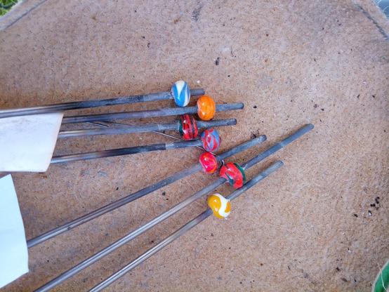 7 Stahldorne mit bereits abgekühlten, farbigen Glasperlen liegen auf einer Lederunterlage in wunderschönen Farben: blauweiß, orange, grünrot, blaurot, rot, gelbweiß.