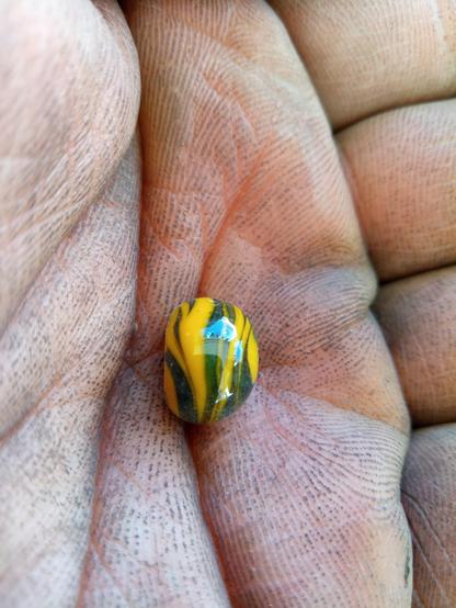 Eine rußige Hand hält eine orangegrün verschleierte Glasperle, in der sich blauer Himmel spiegelt.