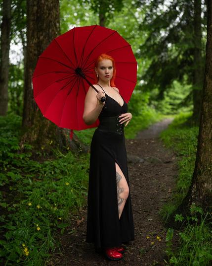 Red-haired person holding a red umbrella, wearing a black dress with a thigh-high slit revealing a tattooed leg, and red shoes. Standing on a forest path flanked by tall trees and greenery.