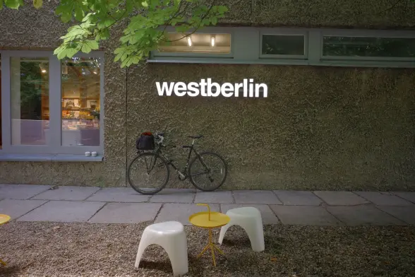 The Surly Long Haul Trucker leaning against a wall with the lit up logo of the coffee shop with the name westberlin above. In front a small outdoor table with low chairs in the pebble garden.