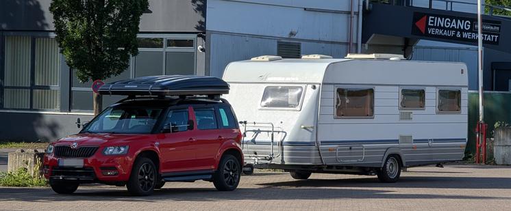 Ein rotes Auto - Skoda Yeti - mit einem angehängten Wohnwagen steht auf einer gepflasterten Fläche vor einem Gebäude mit einem Schriftzug an einer Wand. Auf dem Dach des Zugfahrzeugs ist ein Dachzelt montiert.