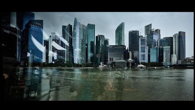 A view of a modern city skyline with tall skyscrapers. The buildings are reflected in a body of water in the foreground. The scene includes a cloudy sky, giving the image a moody atmosphere
