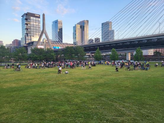 Shot of a group of several hundred cyclists gathered in Paul Revere Park in Boston, Massachusetts. The Zakim bridge and Charles River are visible in the background