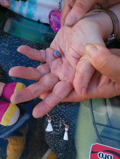 Die Hand einer älteren Person wird von zwei anderen Händen so geführt, dass eine orangeblaue Perle darauf liegen bleibt.