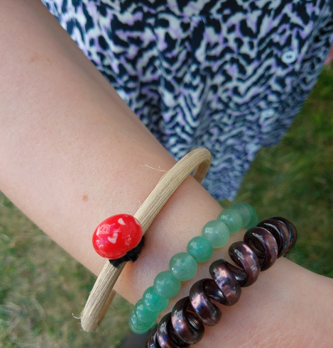 Ein Unterarm mit drei Schmuckringen. Eine dunkelviolette Plastikspirale, eine grüne Plastikperlenschnur und ein Rattanreifen, an dem eine rote Glasperle mit weißen Punkten befestigt ist.
